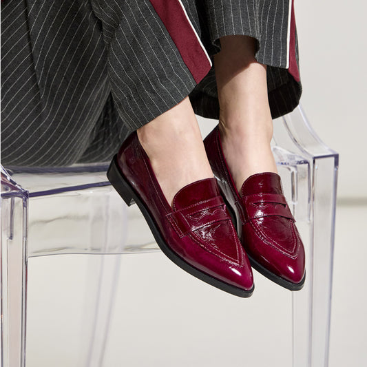 A person wearing glossy deep burgundy patent leather penny loafers with pointed toes, seated on a clear acrylic chair. The outfit includes dark gray pinstriped trousers with red side stripes.