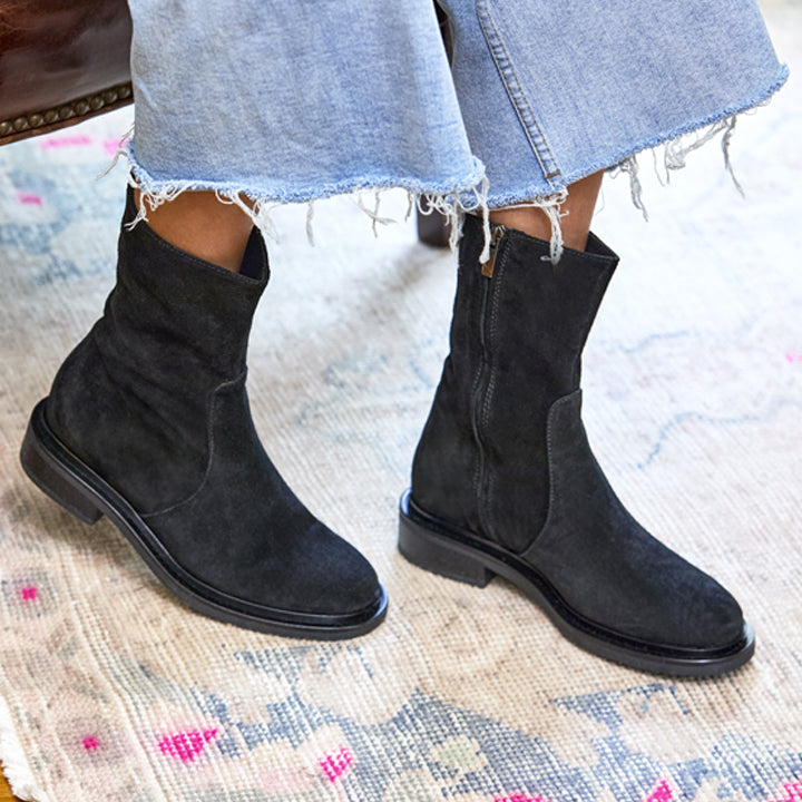 Black suede boots worn with light blue jeans on a patterned rug.