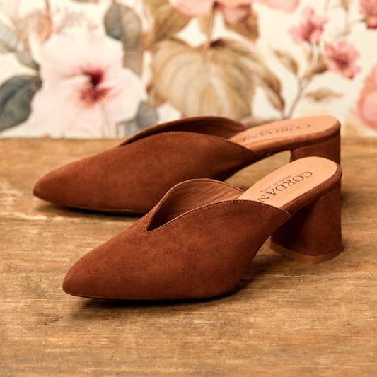 A pair of brown suede mule heels with block toes and low block heels, displayed on a rustic wooden surface with a floral wallpaper background. The brand name 'Cordani' is visible on the insole.