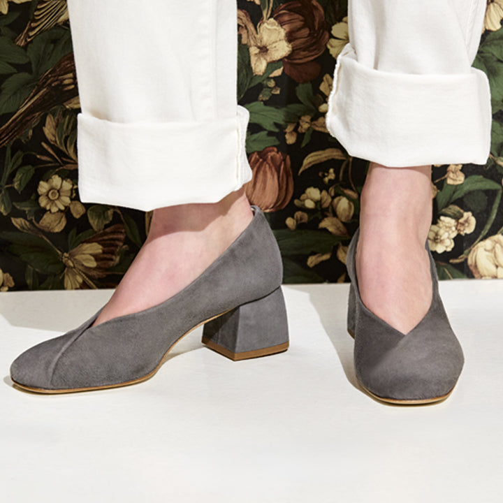 A person wearing gray suede block-heel pumps with a V-shaped toe, paired with white cuffed pants, standing against a dark floral wallpaper background.