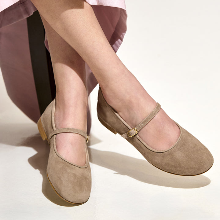 A pair of taupe suede Mary Jane flats with low block heels, shown on a model's feet against a clean off-white background.