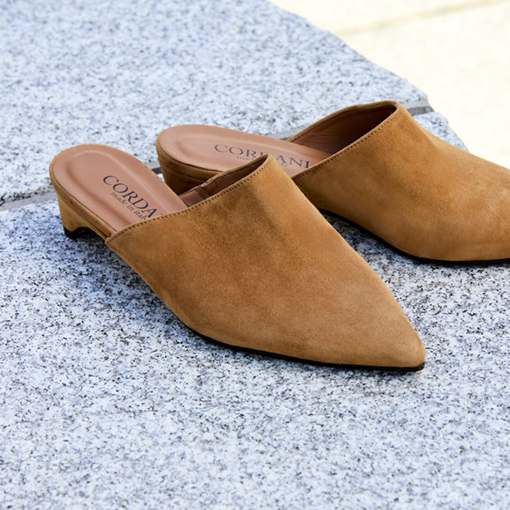A pair of tan suede mule slip-on shoes with low block heels, resting on a gray stone surface. The brand name 'CORDANI' is printed on the insole.