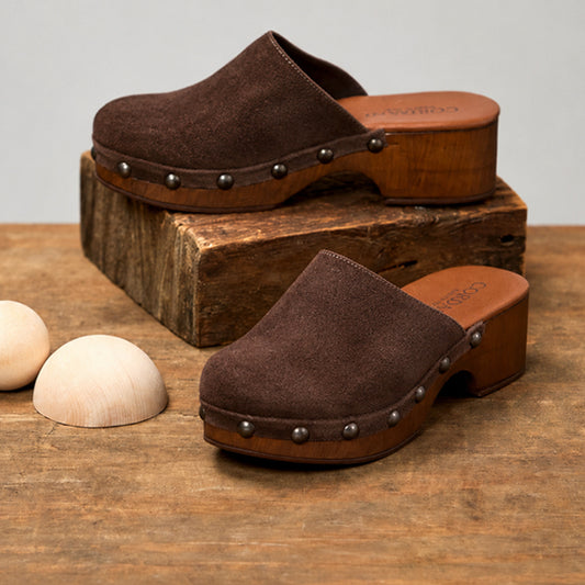 A pair of dark brown suede clogs with wooden platform soles and metal studs, displayed on a rustic wooden block and tabletop with two small wooden beads.