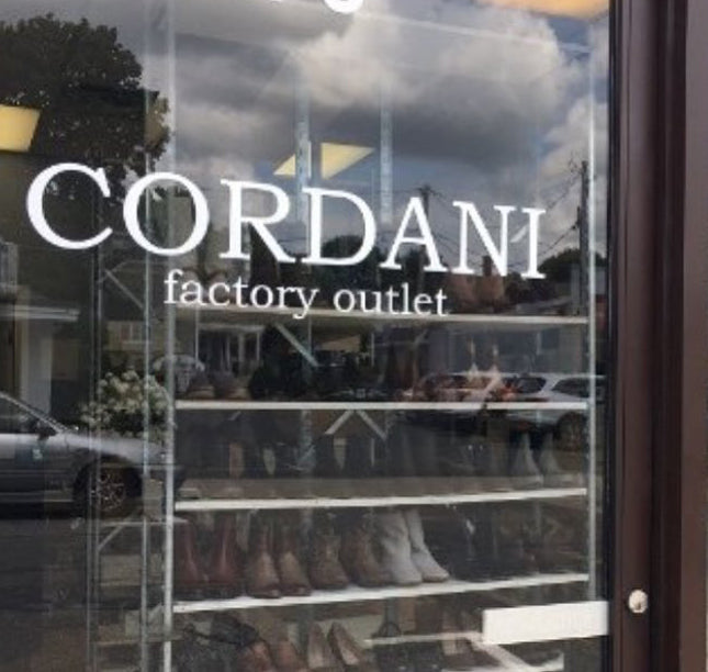 Exterior view of the Cordani Factory Outlet storefront window with white lettering on the glass and shelves of boots visible inside.