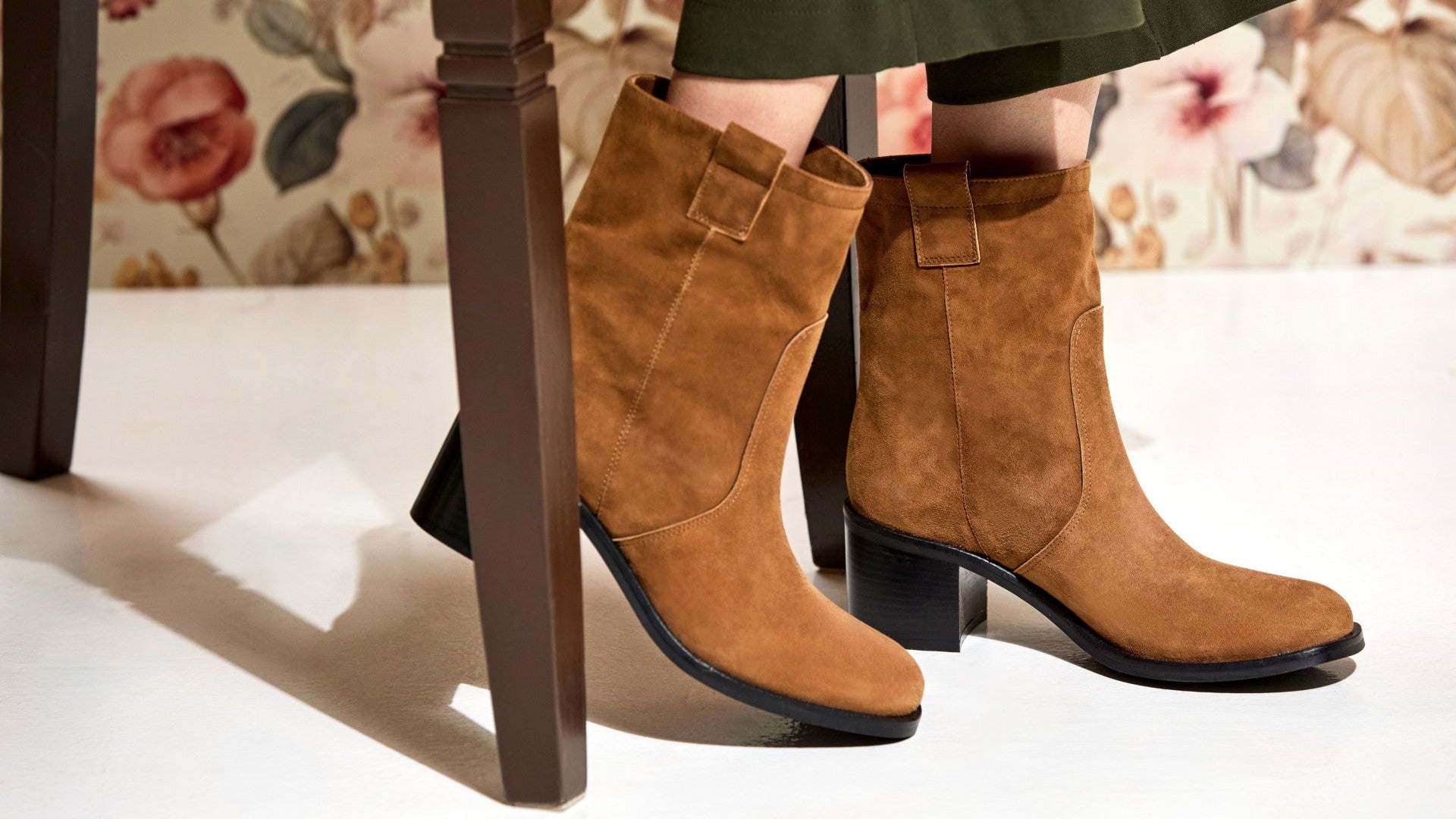 A pair of tan suede ankle boots with black block heels, shown on a model's feet next to a dark wooden chair leg in a styled indoor setting.