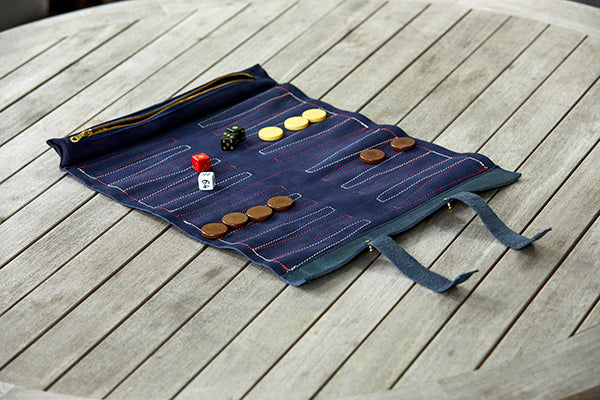 A travel backgammon set rolled up on a wooden table, with game pieces and leather trim visible.
