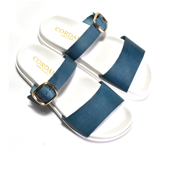White sandals with blue straps and gold buckles on a white background, featuring the brand 'Corda'.
