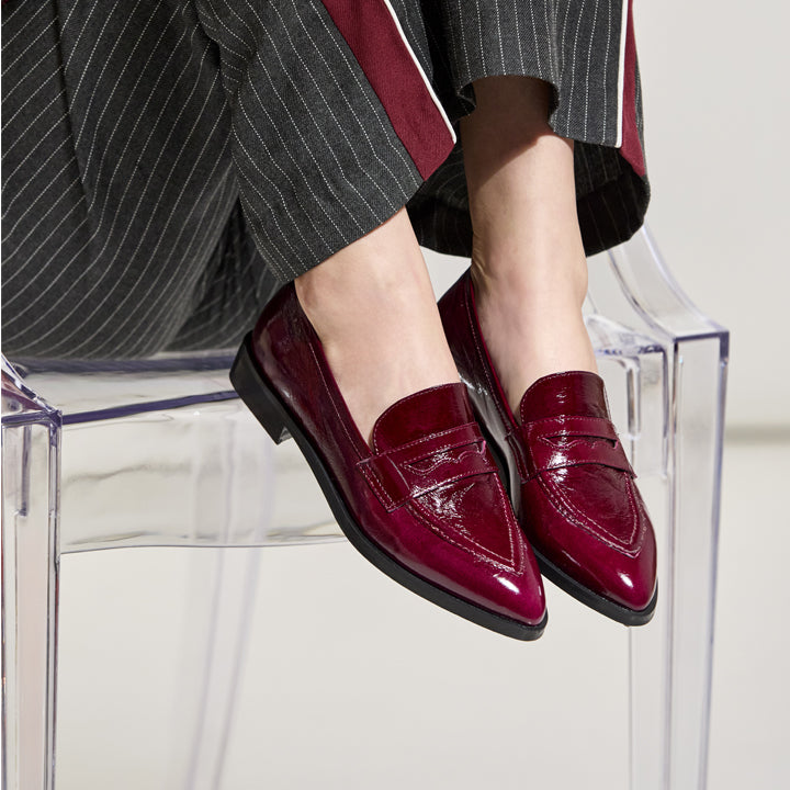 A person wearing glossy deep burgundy patent leather penny loafers with pointed toes, seated on a clear acrylic chair. The outfit includes dark gray pinstriped trousers with red side stripes.