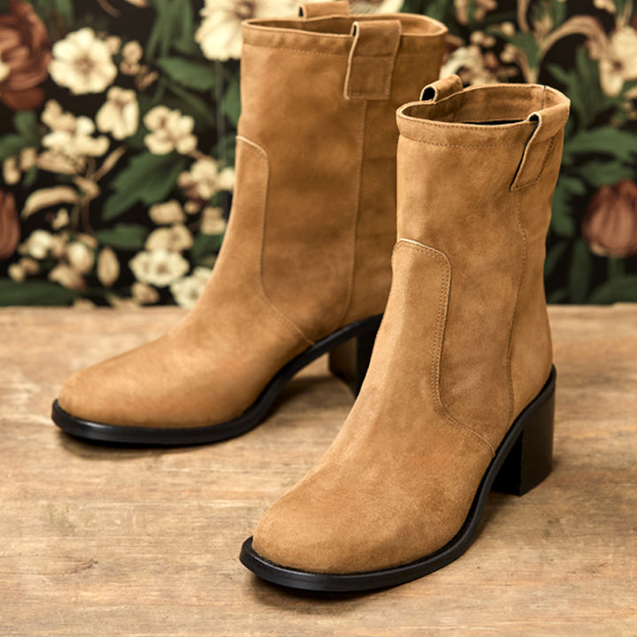 A pair of tan suede ankle boots with block heels, displayed on a rustic wooden surface with a floral wallpaper background.