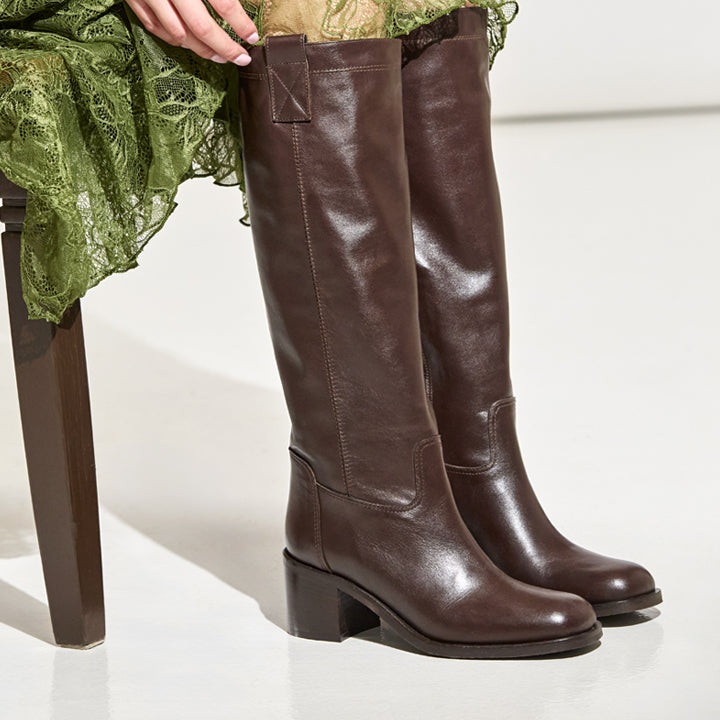 A pair of dark brown knee-high leather boots with block heels, shown on a model seated on a chair wearing an olive green skirt.