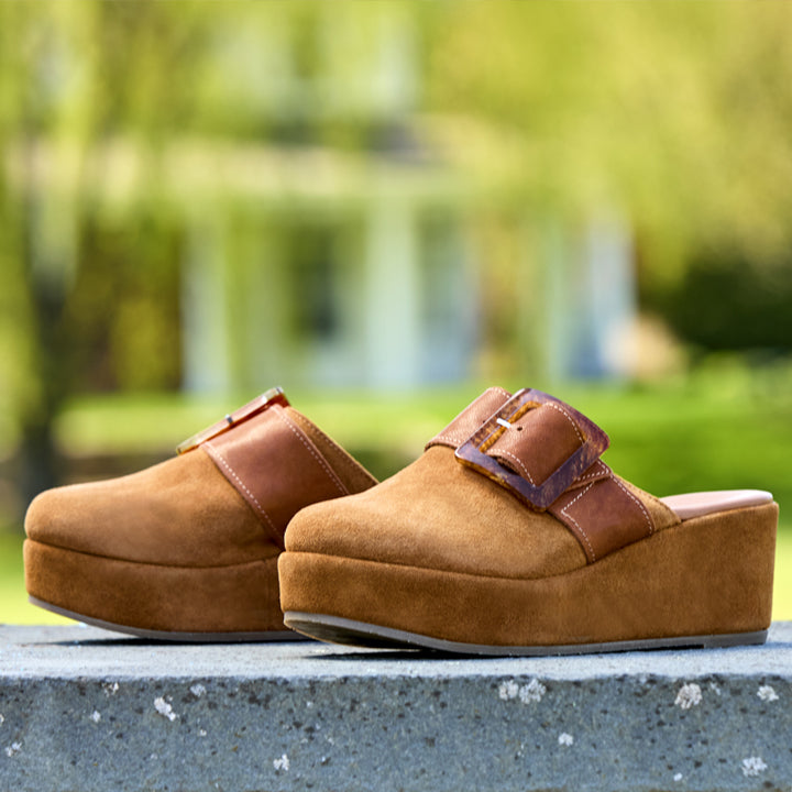A pair of brown platform clogs with wide brown leather straps and decorative buckles, resting on a gray stone ledge with a blurred green garden background.