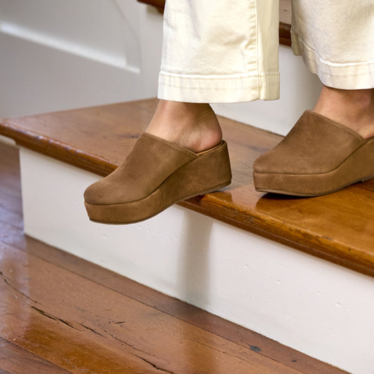 Person wearing brown suede clogs climbing wooden stairs.