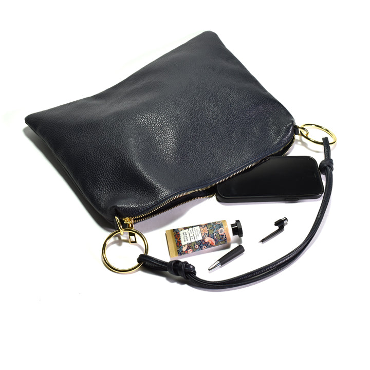 A navy pebbled leather clutch pouch with gold-tone hardware, shown open with its contents arranged below on a white background. The pouch includes a wrist strap and zipper, and is displayed with a black phone, a small patterned card, and two black accessories.