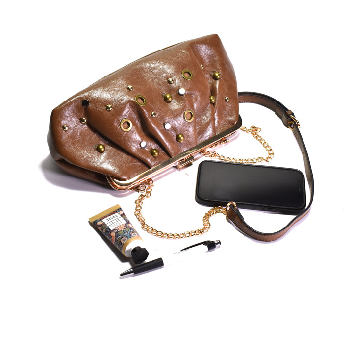 A brown leather clutch handbag with gold-tone hardware, studs, and a chain strap, shown on a white background with a black phone, a tube of hand cream, and a lip gloss arranged beside it.