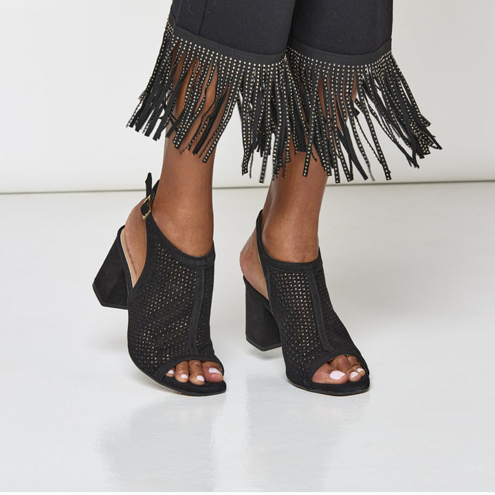Black high-heeled shoes with open-toe design worn with black pants featuring fringe and embellishments.