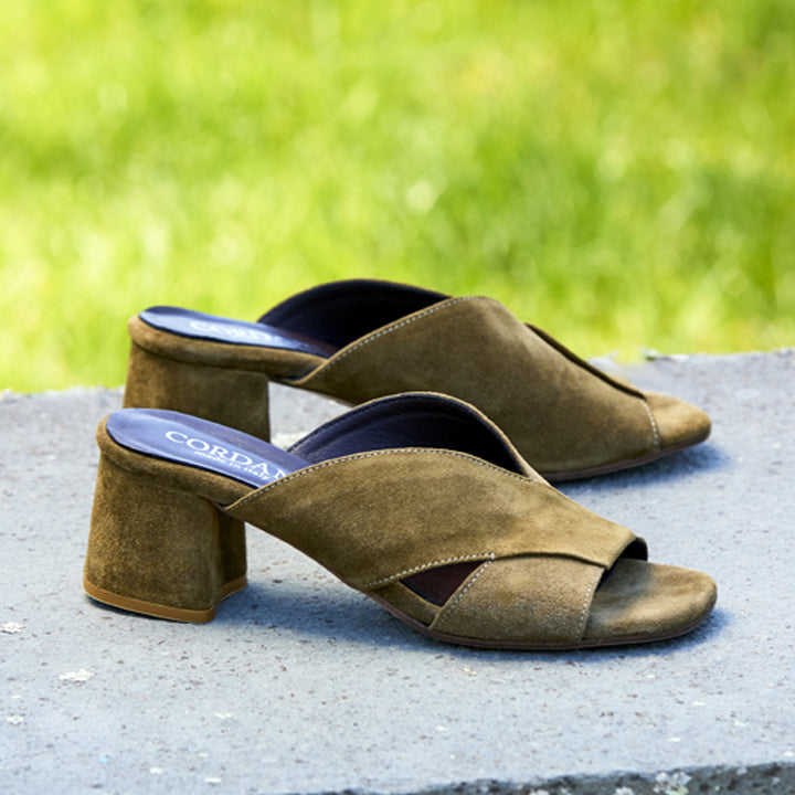 A pair of women's heeled mule sandals in olive-brown suede, shown outdoors on a concrete surface with a blurred green grass background. The brand name 'CORDANI' is visible on the insole.