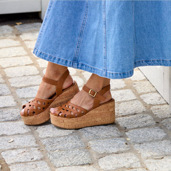 Brown cork wedge sandals worn with a blue skirt on a stone pavement.