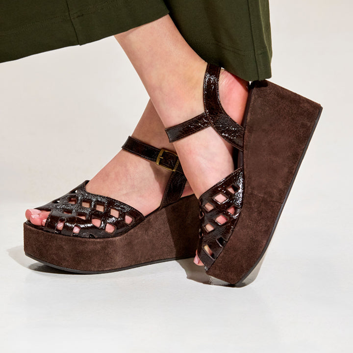 A pair of women's brown platform wedge sandals with cutout lattice detailing on the toe and straps, shown on a model's feet against a light gray studio background.