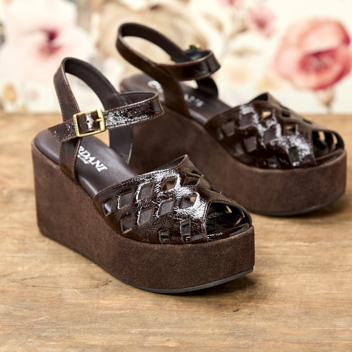 A pair of dark brown platform wedge sandals with cutout detailing on the toe and upper straps, featuring a glossy patent finish and a gold buckle, displayed on a wooden surface with a blurred floral background.