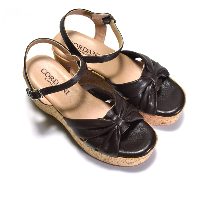 A pair of women's wedge sandals by Cordani, featuring dark brown leather straps with a bow detail, adjustable ankle straps, and cork-wrapped wedge heels, shown on a white background.