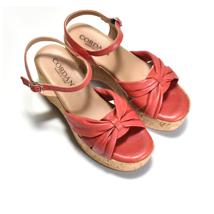 A pair of red leather wedge sandals with ankle straps, decorative crisscross bow details on the toes, and cork-wrapped wedge heels. The brand name 'CORDANI' is visible on the tan insoles.