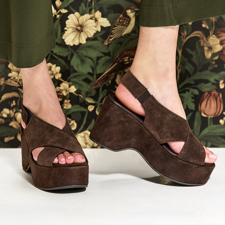 A pair of dark brown suede platform wedge sandals with cutout toes and ankle straps, shown on a model's feet against a dark floral background.