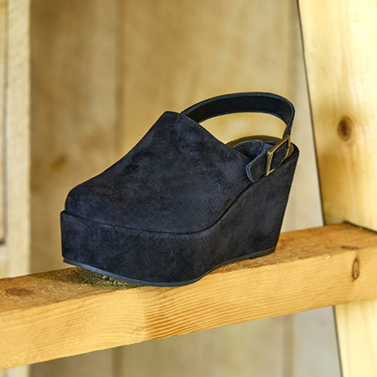 Black suede wedge clog on a wooden shelf with a neutral background