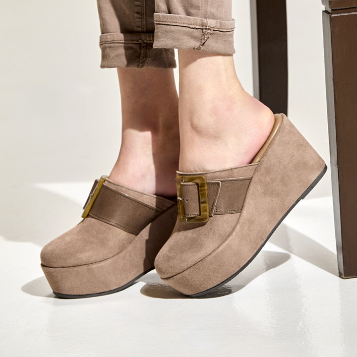 A person wearing taupe-brown platform mule sandals with a wide strap and gold-tone buckle, paired with rolled-up brown pants, shown against a clean white studio background.