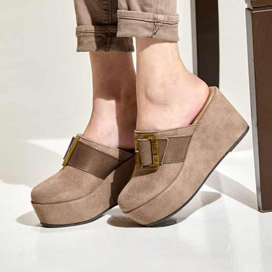A person wearing taupe-brown platform mule sandals with a wide strap and gold-tone buckle, paired with rolled-up brown pants, shown against a clean white studio background.