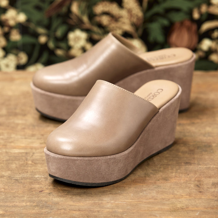 A pair of taupe platform mule sandals with smooth leather uppers and suede wedge soles, displayed on a rustic wooden surface with a blurred background of green foliage and dried flowers.