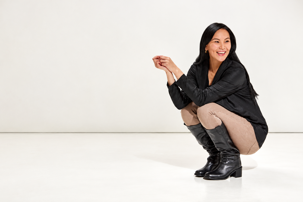 A smiling woman crouching in a studio setting, wearing a black long-sleeve top, beige leggings, and black knee-high boots against a clean white background.