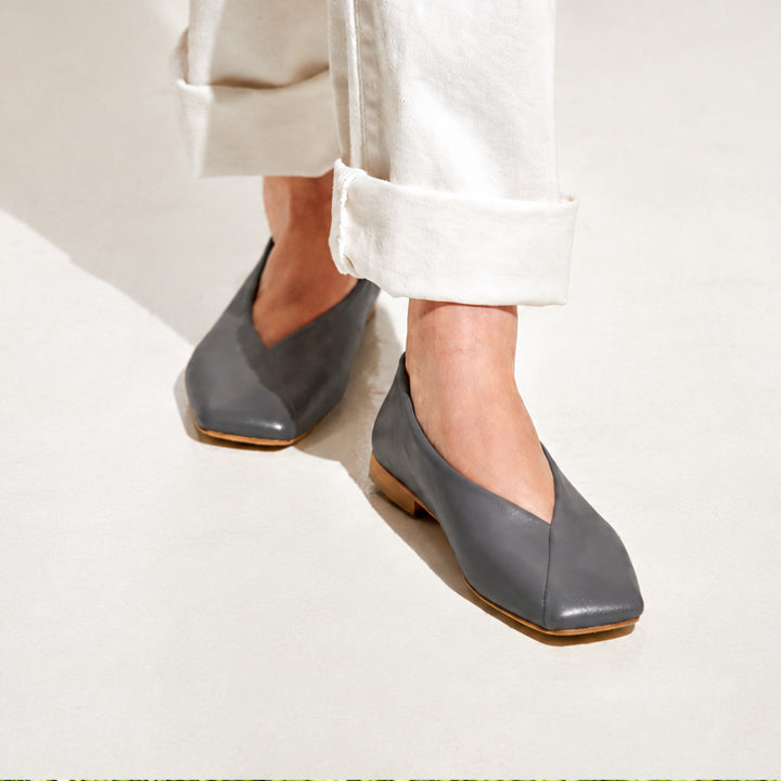 A person wearing gray leather square-toe flats with rolled-up white pants, standing on a light beige floor under bright natural sunlight.