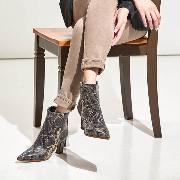 A person seated on a wooden chair wearing pointed-toe ankle boots with a snakeskin pattern in gray, beige, and black. The person is also wearing tan cuffed pants and a dark top, photographed in a bright studio setting.