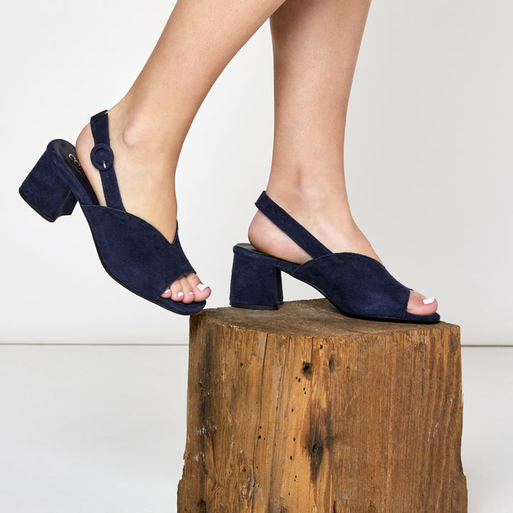 Navy blue high-heeled sandals worn by a person on a wooden block against a white background