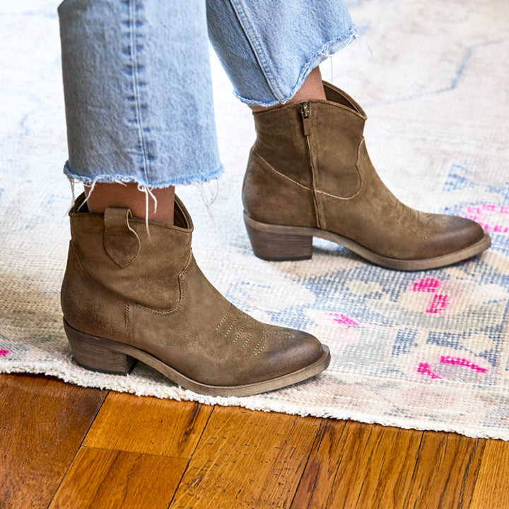 Brown ankle boots worn with light blue jeans on a wooden floor.