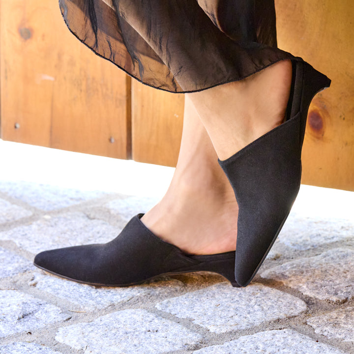 A person wearing black pointed-toe slip-on wedge shoes with sheer black fabric draped at the ankles, standing on a stone surface in front of a wooden wall.