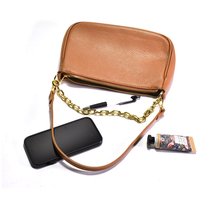 Brown leather handbag with gold chain strap, black phone, pen, and tube of lotion on a white background