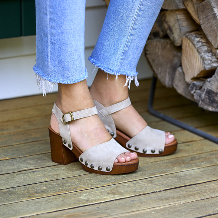 Beige suede sandals with wooden soles worn by a person on a wooden deck.