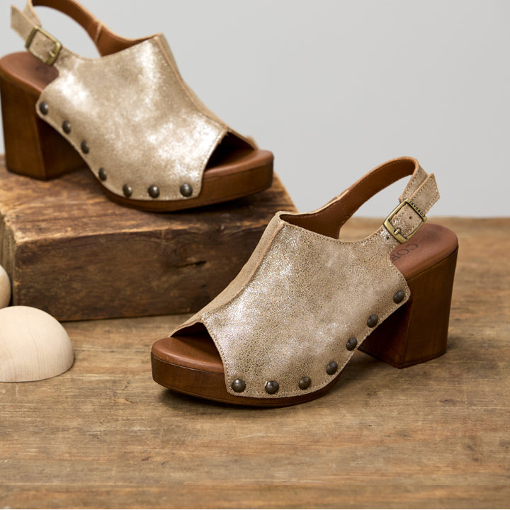 A pair of women's open-toe block-heel sandals with metallic champagne-colored straps and dark metal studs, displayed on a rustic wooden surface with one shoe on a wooden block.
