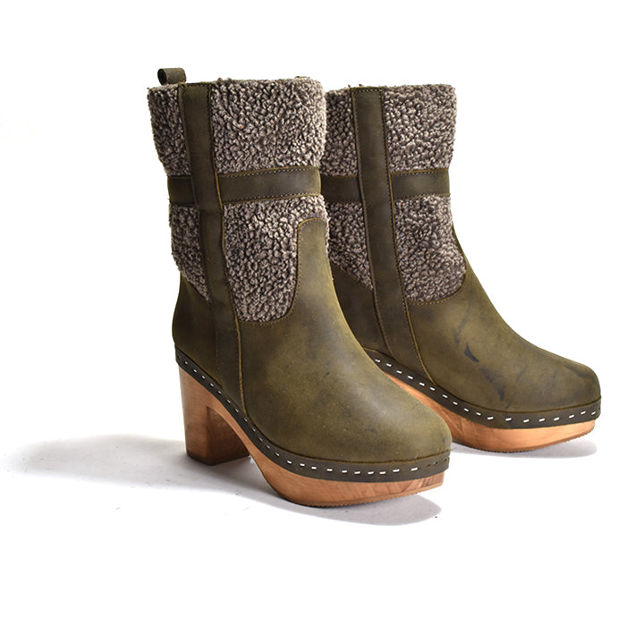 A pair of women's mid-calf boots in distressed olive green leather with brown faux-shearling panels on the sides and backs, featuring wooden block heels and staples around the sole.